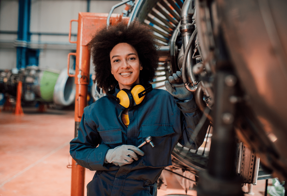 An image of a woman wearing blue overalls stands confidently next to a large jet engine, carefully working on repairs with her precision engineering tools. 